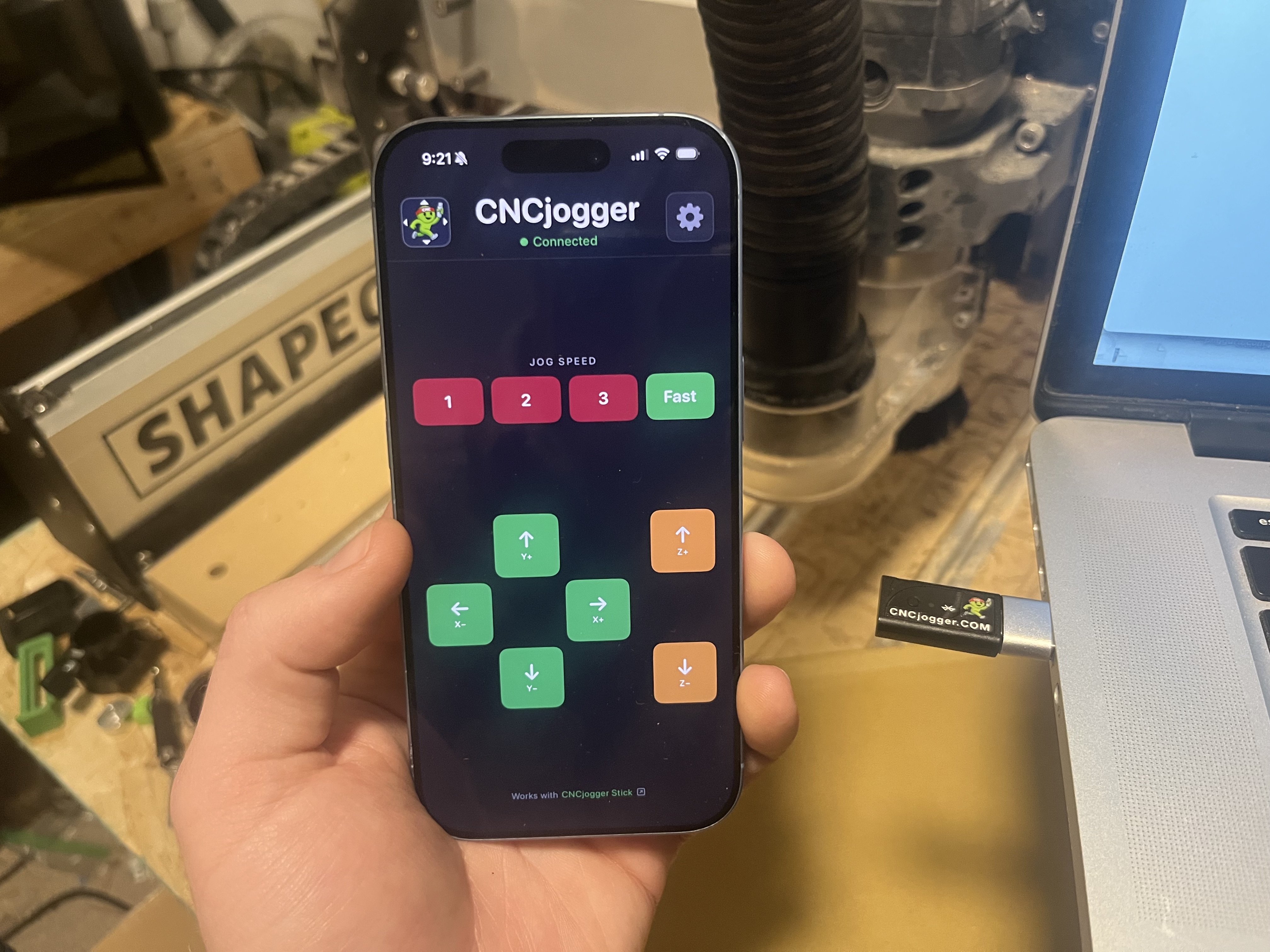 CNCjogger — iPhone app showing wireless jog controls with USB-C receiver plugged into laptop, Shapeoko CNC in background